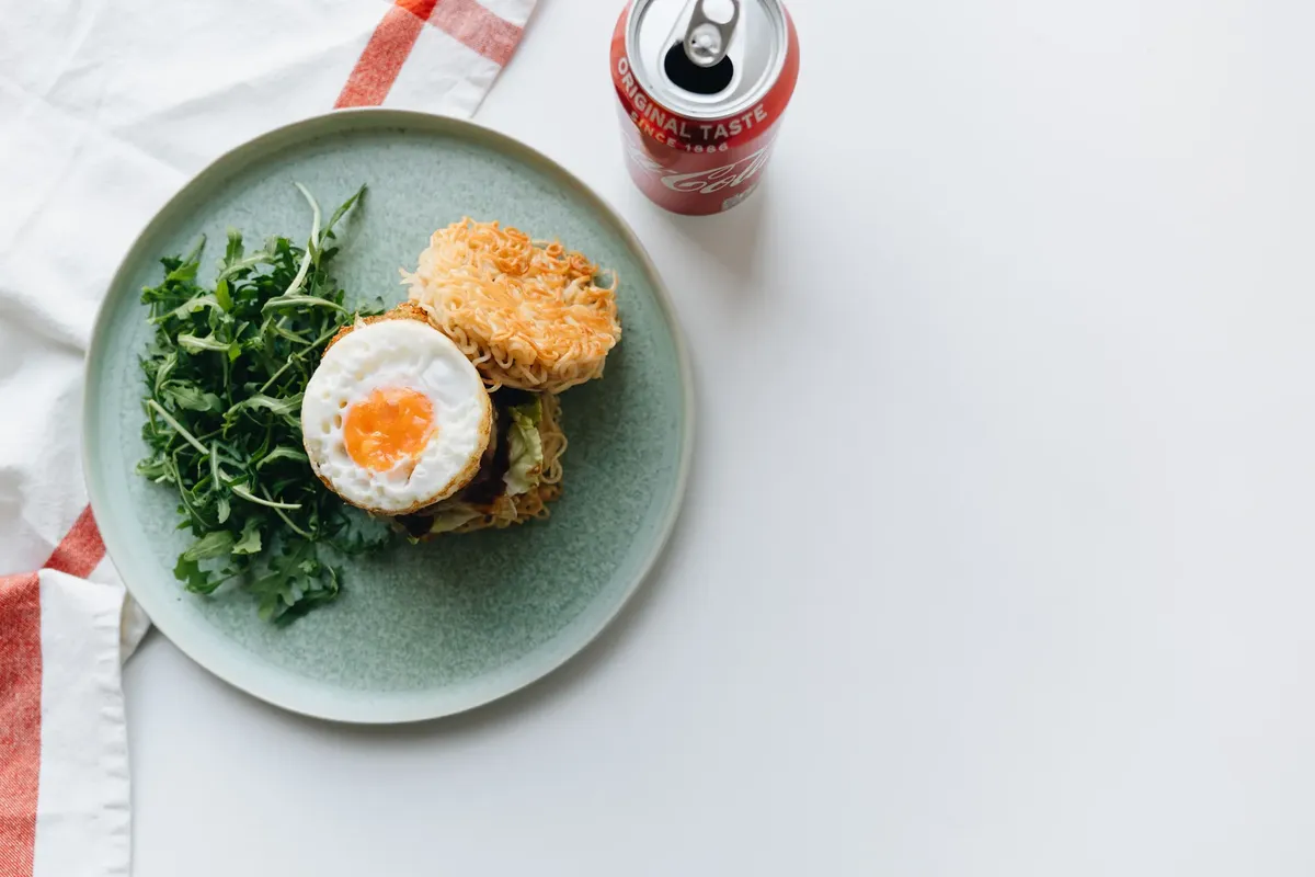 Gourmet ramen burger topped with egg and greens alongside a refreshing soda can.