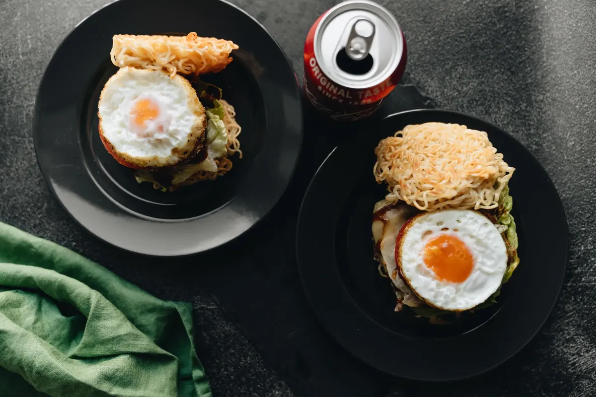 Top view of ramen burgers with fried eggs and a soda can offering a delicious twist on classic snacks.