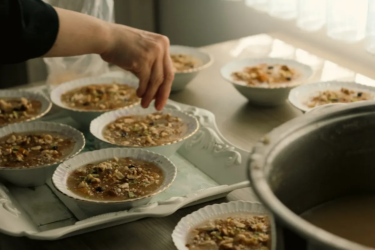 A person garnishes bowls of traditional dessert with nuts and spices on a kitchen counter.