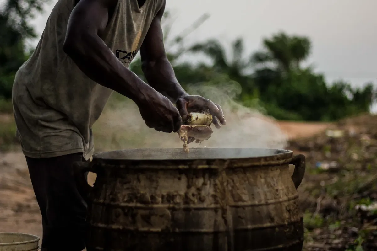 An African man cooking food in a large pot outdoors in a rural Nigerian village.