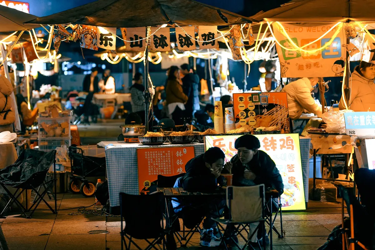 Bustling night market scene with food stalls illuminated and patrons enjoying street food under warm lights.